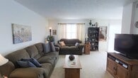 This two-floor home offers a warm and inviting atmosphere with a thoughtfully arranged interior. The living room serves as the heart of the main level, featuring wall-to-wall carpeting in a soft neutral tone that adds a sense of comfort underfoot. Two generously sized sofas in earthy brown tones are arranged to encourage conversation, complemented by a wooden coffee table at the center. Natural light filters through a window dressed with elegant cream curtains hung on a classic rod, brightening the room with a soft, airy glow. A tall floor lamp and a table lamp add layered lighting options for the evening hours. Along one wall, a dark wood bookcase displays a charming collection of figurines and decorative items, lending the room a personal and curated character. Framed artwork adorns the walls, contributing to the home's warm, lived-in aesthetic. A lush potted plant in the corner introduces a touch of nature to the space. The textured ceiling adds subtle visual interest overhead. The home's two-floor layout provides ample space for both daily living and private retreats, making it a well-rounded and functional residence suited for a variety of lifestyles.