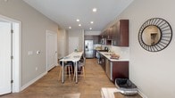 This single-floor residence at REVA Main St. in Rahway, NJ offers a thoughtfully designed open-concept layout that maximizes functionality and comfort. Rich hardwood-style flooring runs throughout the home, creating a cohesive and contemporary aesthetic.

The living area is bright and spacious, benefiting from large windows that invite generous natural light and provide outdoor views. Recessed lighting and clean neutral walls enhance the airy feel of the space, while a sliding glass door connects the living area to an outdoor balcony.

The kitchen is a standout feature, showcasing rich dark cabinetry contrasted by light quartz countertops and a classic white subway tile backsplash. A full suite of stainless steel appliances—including a gas range, microwave, refrigerator, and dishwasher—provides modern convenience. A breakfast bar with bar seating adds flexibility for casual dining.

The bedroom is a calm, well-lit retreat featuring large windows, a ceiling fan, and softly textured walls that lend a serene ambiance. A second bedroom configuration is also visible, offering similar natural light and a clean, neutral palette.

The bathroom is finished with dark cabinetry, a light countertop vanity, and hardwood-style flooring, maintaining the consistent modern design found throughout the home.