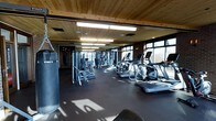This fitness facility offers a thoughtfully designed workout environment that blends rustic and industrial aesthetics. The space is characterized by warm wood-paneled ceilings and exposed brick walls, creating an inviting atmosphere that sets it apart from a typical gym. Large windows along one side of the facility flood the space with natural light and provide views of the surrounding outdoor scenery, enhancing the overall workout experience.

The expansive workout floor is finished with durable dark rubber flooring, providing a safe and functional surface for a wide range of exercises. The facility is well-equipped with a comprehensive selection of cardio machines, including treadmills and ellipticals, neatly arranged along the window-facing wall to take advantage of the natural light and outdoor views. Strength training equipment, including cable machines and weight stations, are systematically arranged throughout the interior of the space. A heavy punching bag is also present, catering to those interested in boxing or combat fitness training.

Overhead fluorescent lighting supplements the natural light, ensuring the space remains well-lit throughout. The combination of high-end equipment, architectural warmth, and ample natural light makes this a versatile and welcoming fitness destination.