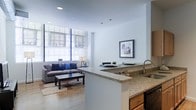 This single-floor space showcases an open-concept layout that integrates the kitchen and living area into one cohesive, free-flowing environment. The kitchen is appointed with granite countertops, warm wood cabinetry, and a peninsula that doubles as a breakfast bar, providing both additional workspace and a natural visual divide between the two zones. A stainless steel sink adds a sleek, modern touch to the culinary space.

The living area benefits from generous natural light streaming through large, floor-to-ceiling windows dressed with sheer roller shades, creating a bright and airy atmosphere. Hardwood flooring extends throughout, lending warmth and continuity to the space. Recessed lighting in the white ceiling complements the natural light, ensuring the interior remains well-illuminated at all hours.

The living area comfortably accommodates a full sofa and coffee table arrangement, grounded by a neutral area rug. Clean white walls serve as a versatile backdrop, with framed artwork adding a refined decorative accent. Overall, the space presents a modern, minimalist aesthetic with a welcoming and functional character.