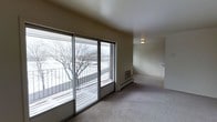 This single-floor space offers a clean, functional layout designed for comfortable everyday living. The living area serves as the focal point of the unit, featuring large sliding glass doors that span much of the exterior wall, flooding the room with natural light and providing direct access to a private balcony. The balcony overlooks an open outdoor setting with views of mature trees, making it a standout feature of the space. The living area is finished with wall-to-wall carpeting in a neutral tone, contributing to a warm and understated ambiance. A wall-mounted heating unit is conveniently positioned near the sliding doors, ensuring efficient climate control. The ceiling is smooth and finished in a crisp off-white, complementing the light-toned walls throughout. A ceiling-mounted light fixture provides additional illumination. The open layout allows for flexible furniture arrangements, and a connecting passage leads to adjacent areas of the unit. Overall, the space presents a straightforward and practical design with an emphasis on natural light, outdoor connectivity, and a neutral color palette that serves as a versatile backdrop for a variety of interior styles.
