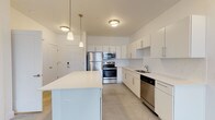 This single-floor unit showcases a clean, contemporary design throughout its well-organized layout. The kitchen serves as the centerpiece of the space, featuring crisp white cabinetry with slim bar-pull hardware, a white quartz-topped center island, and a full suite of stainless steel appliances including a refrigerator, range, microwave, and dishwasher. Pendant lighting with warm-toned glass shades hangs above the island, complementing the flush ceiling fixtures overhead. White subway tile backsplash lines the walls, reinforcing the kitchen's bright, minimalist aesthetic. Polished concrete floors extend throughout the main living and kitchen areas, lending an industrial-modern character to the space.

The bathroom continues the unit's neutral palette with white walls, a tiled shower-tub combination, a floating vanity, and concrete flooring. Brushed nickel hardware and an assist bar add functional detail. The bedroom offers a quieter, softer environment with plush neutral carpeting underfoot and a window fitted with horizontal blinds that allows natural light to filter in. The overall design of this unit is cohesive and understated, prioritizing clean lines, functional layout, and a bright, open ambiance throughout.