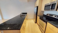 This single-floor space showcases a thoughtfully designed kitchen that blends modern aesthetics with functional layout. The kitchen features striking dark granite countertops that contrast elegantly with the light natural wood cabinetry, which is fitted with sleek bar-pull hardware for a contemporary look. The countertops extend generously along both sides of the galley-style kitchen, providing ample prep and workspace. A stainless steel undermount sink is seamlessly integrated into the counter, complemented by a modern arc-neck faucet. The full suite of stainless steel appliances — including a French door refrigerator, electric range with oven, and built-in microwave — adds a polished, professional-grade feel to the space. Light hardwood flooring runs throughout, lending warmth and a sense of continuity that brightens the interior. The cabinetry extends to the upper walls, maximizing storage capacity, while the clean white walls keep the overall atmosphere open and airy. The layout is efficient and well-organized, making excellent use of the available space to deliver a kitchen that is both visually appealing and highly practical for everyday use.