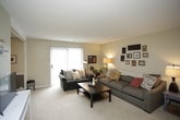 This single-floor residence offers a thoughtfully organized layout with warm, neutral tones and carpet flooring throughout the main living areas. The living room is generously sized, providing ample space for multiple seating arrangements. It receives natural light through a large window fitted with vertical blinds, and wall-mounted shelving adds a practical decorative touch. The space flows naturally into a defined dining area, which features a ceiling fan with a light fixture, a structural beam accent, and a window that brings in additional natural light.

The kitchen is efficiently laid out with white cabinetry extending to the ceiling for abundant storage, light countertops, a stainless steel sink, and white appliances including a range and full-size refrigerator. Wood-look flooring in this area provides a clean contrast to the carpeted rooms.

The primary bedroom is a comfortable retreat with a ceiling fan, soft carpet, and direct access to an en-suite bathroom. A secondary bedroom offers carpet flooring, a window with sheer curtains, and space for a dedicated desk area. The full bathroom features white tile wainscoting, a mirrored medicine cabinet, a sink with a vanity, and a shower-tub combination.