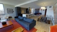 This single-floor space showcases a thoughtfully designed open-plan layout that effortlessly connects the living, dining, and kitchen areas into one cohesive environment. The living area is anchored by a large, deep-blue sofa set atop a bold red area rug, creating a striking focal point against the warm tones of the light hardwood flooring that runs throughout the space. Adjacent to the living area, a substantial wooden dining table paired with a matching bench offers generous seating and an inviting atmosphere for gatherings. The kitchen is a standout feature, boasting sleek dark cabinetry, integrated appliances including a built-in oven and a full-height refrigerator, and a large central island topped with a white countertop. The island is lined with a row of modern bar stools, making it a functional and social hub. A stainless steel pendant hood serves as an industrial accent above the cooktop. Recessed lighting throughout ensures the space is well-illuminated, complemented by a large pendant light above the dining area. Further into the space, a defined lounge nook with built-in shelving and colorful accent chairs adds a relaxed, curated corner. The overall ambiance is modern and refined, with a harmonious blend of dark, light, and warm tones.