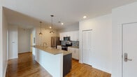 This single-floor unit offers a clean, contemporary design with a thoughtful layout that balances open communal spaces and private rooms. The heart of the home is a well-appointed kitchen featuring white shaker cabinetry, quartz countertops, a mosaic tile backsplash, and a full suite of stainless steel appliances including a French door refrigerator, electric range, and built-in microwave. A large island with a farmhouse-style sink and dark wood base provides additional prep space and seating, accented by stylish pendant lighting overhead. The kitchen flows seamlessly into a bright, open living area finished with warm hardwood flooring and recessed lighting. Sliding glass doors bring in natural light and offer views of the surroundings. The bedrooms are carpeted with a patterned tile-style carpet and are served by windows fitted with blinds. The bathroom is finished with a dark wood vanity, white countertop, large mirror, and brushed nickel fixtures, complemented by light wood-look tile flooring and accessibility grab bars. A stacked in-unit washer and dryer provides added convenience. Throughout the unit, crisp white walls, clean lines, and modern finishes create a bright and cohesive living environment.