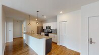 This single-floor unit offers a clean, contemporary design with a thoughtful layout that balances open communal spaces and private rooms. The heart of the home is a well-appointed kitchen featuring white shaker cabinetry, quartz countertops, a mosaic tile backsplash, and a full suite of stainless steel appliances including a French door refrigerator, electric range, and built-in microwave. A large island with a farmhouse-style sink and dark wood base provides additional prep space and seating, accented by stylish pendant lighting overhead. The kitchen flows seamlessly into a bright, open living area finished with warm hardwood flooring and recessed lighting. Sliding glass doors bring in natural light and offer views of the surroundings. The bedrooms are carpeted with a patterned tile-style carpet and are served by windows fitted with blinds. The bathroom is finished with a dark wood vanity, white countertop, large mirror, and brushed nickel fixtures, complemented by light wood-look tile flooring and accessibility grab bars. A stacked in-unit washer and dryer provides added convenience. Throughout the unit, crisp white walls, clean lines, and modern finishes create a bright and cohesive living environment.