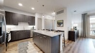This well-appointed 2-bedroom, 2-bathroom residence offers a thoughtfully designed single-level layout with modern finishes and a cohesive aesthetic throughout. The open-concept kitchen and living area serves as the heart of the home, anchored by a large granite-topped island with bar seating and sleek pendant lighting overhead. Dark wood-grain cabinetry, a classic subway tile backsplash, and a full suite of stainless steel appliances — including a french door refrigerator — complete the kitchen. Wood-style flooring flows seamlessly throughout the entire residence, adding warmth and continuity. The living room features access to a private balcony through full-length glass doors, inviting generous natural light. A dedicated dining area sits adjacent, creating a natural flow ideal for entertaining. The spacious primary bedroom includes a walk-in closet and direct access to an en-suite bathroom, which features a granite-topped vanity with dark cabinetry and a full soaking tub with shower. The second bedroom is equally well-sized, also offering a closet and natural light. A hallway connects the bedrooms and living spaces, providing a practical and organized floor plan. Recessed lighting and neutral wall tones contribute to the clean, contemporary ambiance throughout.