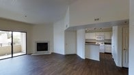 This single-story space showcases an open-concept layout that seamlessly connects the living area and kitchen. The living room benefits from soaring vaulted ceilings that amplify the sense of space, while dark hardwood flooring runs throughout, providing a warm contrast to the bright white walls. A tiled fireplace with a classic surround serves as a focal point in the living area, adding character and a cozy element to the room.

An arched passthrough connects the living space to the kitchen, which is outfitted with white cabinetry, a white appliance suite including a gas range and built-in microwave, and dark countertops. Track lighting illuminates the kitchen workspace efficiently. A ceiling vent ensures proper climate control throughout. The entry door, visible from the living area, is framed neatly within the open floor plan. Overall, the space maintains a clean, neutral palette with practical finishes, creating a functional and inviting single-level environment.