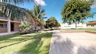 This single-story property is distinguished by its verdant, tropical outdoor environment. The exterior grounds are generously landscaped, featuring tall, mature palm trees and broad-canopied shade trees that frame the property and provide natural cover. A well-maintained concrete driveway and walkway run alongside neatly trimmed green lawns, creating a clean, open approach to the building. A low brick border accent adds a classic detail to the landscaping. The building exterior, visible in the background, presents a light-toned facade with a straightforward, functional architectural style that is common in warm-climate construction. The grounds convey a sense of spaciousness, with ample open lawn area and established plantings that contribute to a calm, shaded atmosphere under open blue skies. The interior images provided do not offer sufficient detail to describe indoor features accurately.