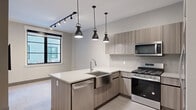 This efficiently designed single-floor unit delivers a modern living experience through carefully selected finishes and a cohesive contemporary aesthetic. The open-concept kitchen serves as the centerpiece of the space, anchored by a large island with a white quartz countertop and an undermount sink. Wood-toned flat-panel cabinetry lines the walls, complemented by a full suite of stainless steel appliances including a gas range, over-the-range microwave, refrigerator, and dishwasher. Three industrial-style pendant lights hang above the island, adding character and focused task lighting, while track lighting provides additional illumination throughout. Light-toned luxury vinyl flooring runs throughout the space, enhancing the bright, airy feel. A window allows natural light to filter into the kitchen and living area, which also features a built-in media niche. The bathroom maintains the same modern design language, with a wood-grain vanity, white quartz countertop, open wall-mounted shelving, and a glass-enclosed shower. An in-unit washer is also accommodated within the bathroom area. The overall ambiance is sleek, minimal, and functional, making efficient use of every square foot.