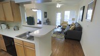 This single-floor space showcases an open-concept layout that seamlessly connects the kitchen and living area. The kitchen features a wraparound breakfast bar with a white countertop and an integrated sink, complemented by warm honey-toned wood cabinetry. A pendant light fixture hangs above the bar, providing focused task lighting. The kitchen opens directly into the living area, creating a connected, social atmosphere ideal for entertaining.

The living area is finished with rich hardwood flooring and benefits from abundant natural light streaming through multiple windows, including a set of glass-paneled doors that brighten the space considerably. A ceiling fan is centrally positioned overhead, adding both function and a casual aesthetic. The room comfortably accommodates a full seating arrangement, with ample space for a sectional sofa, accent chair, coffee table, and a patterned area rug that anchors the layout. A pass-through window visible from the kitchen connects to an additional room, likely a bedroom, further enhancing the open and airy feel of the overall floor plan.