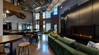 This expansive single-floor communal space masterfully blends industrial and contemporary design elements to create a dynamic, inviting environment. Soaring double-height ceilings anchor the main lounge area, where exposed ductwork, a distinctive slatted ceiling fixture with integrated lighting, and pendant lights contribute to a raw yet refined aesthetic. Original brick walls rise dramatically toward the upper windows, flooding the interior with natural light and reinforcing the building's industrial heritage.

Polished concrete floors stretch throughout, providing a sleek, reflective surface that ties the various zones together. The centerpiece of the main lounge is a floor-to-ceiling matte black feature wall housing a linear gas fireplace, flanked by a plush forest-green velvet sectional sofa adorned with patterned throw pillows. Nearby, a more intimate seating area features a low-profile dark wood coffee table, a leather lounge chair, and velvet accent chairs positioned beneath tall windows overlooking an outdoor courtyard.

A café-style social hub offers bar-height tables and industrial stools, alongside a workstation counter beneath a mezzanine level. A billiards table adds a recreational element to the space. The entry corridor showcases vibrant neon-inspired artwork against polished walls, setting an energetic tone. Throughout, bold sculptural art pieces, including an organic wall installation, reinforce the space's curated, gallery-like character.