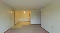 This single-floor space features an open-concept layout that seamlessly connects the main living area to an adjoining room, likely a dining or kitchen space. The living area is finished with wall-to-wall carpet, providing a comfortable and warm underfoot feel, while the connected space transitions to a light-toned tile floor with a subtle pattern. A ceiling fan is installed in the adjoining area, offering both lighting and air circulation. The walls throughout are painted in a neutral white tone, contributing to a bright and clean aesthetic. An HVAC vent is visible near the ceiling, indicating a central climate control system. The space includes standard baseboards along the perimeter and features a doorway on the left side, likely leading to additional rooms or an entry point. An arched or recessed passageway on the right side adds a subtle architectural detail, hinting at further rooms beyond. The overall layout is straightforward and functional, offering a practical flow between spaces suited for everyday living.
