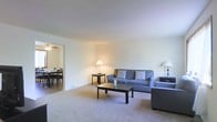 This two-floor space offers a clean, functional layout with a welcoming living area as a central feature. The living room is generously sized, finished with wall-to-wall carpeting and neutral-toned walls that create a bright, understated atmosphere. It is furnished with a pair of grey sofas arranged around a dark-wood coffee table, complemented by a matching side table and floor lamp that add warmth to the room. The space flows openly into an adjacent dining area, visible through a wide doorway. The dining room features a ceiling fan with integrated lighting and accommodates a full dining table with seating for multiple guests. The open-concept connection between these two rooms enhances the sense of spaciousness and makes the layout well-suited for both everyday living and entertaining. The overall aesthetic throughout is simple and neutral, with light walls and carpet flooring maintaining a cohesive, understated look. With two floors, this space provides ample room across multiple levels, offering flexibility in how the layout can be utilized.