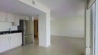 This single-floor space is characterized by a clean, modern aesthetic with an open-concept layout that promotes a sense of spaciousness. Large-format light gray ceramic tiles span the entire floor, providing a cohesive and polished look throughout. The interior is defined by crisp white walls and ceilings, lending a bright and airy atmosphere to the space.

The kitchen area is positioned to one side and features white upper cabinetry and a stainless steel French-door refrigerator, contributing a sleek, contemporary feel. A structural column with a clean white finish serves as a natural visual divider between the kitchen and the open living area, maintaining an unobstructed flow without fully separating the two zones.

The expansive living area benefits from the minimalist design approach, allowing natural light to bounce off the neutral surfaces and enhance the sense of openness. A hallway visible in the background suggests additional rooms beyond the main living space. Recessed ventilation fixtures are integrated into the ceiling, maintaining the clean lines of the overall design. The space presents a well-maintained, move-in ready environment with a neutral palette suitable for a variety of interior styles.