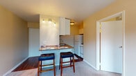 Located at 1428 King Ave in Columbus, Ohio's Clintonville neighborhood, this single-floor unit offers a well-organized and functional layout. The kitchen is a standout feature, designed with white cabinetry, a granite-style countertop, and a peninsula breakfast bar with seating for two — ideal for casual dining. A track lighting fixture illuminates the workspace, while a full suite of white appliances, including a refrigerator and gas range, completes the setup. Rich hardwood flooring in the kitchen transitions into plush carpet throughout the living and sleeping areas, adding warmth and comfort underfoot. The living area is generously sized, featuring a large window that fills the room with natural light and a wall-mounted air conditioning unit for climate control. A closet with louvered doors adds convenient storage. The bedroom continues the carpeted aesthetic and includes its own sizable window with a view of the surrounding neighborhood. The bathroom is clean and classic, featuring a full tub-shower combination with white tile surround, a pedestal-style sink, and a mirrored medicine cabinet. Warm, neutral tones throughout the unit create a cohesive and inviting atmosphere from room to room.
