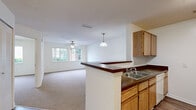 This single-floor, 1-bedroom, 1-bathroom residence offers a practical and comfortable layout designed to maximize every square foot. The open-concept living and dining area is bright and airy, featuring neutral carpet flooring, a ceiling fan, and a large window that fills the space with natural light and garden views. The kitchen is thoughtfully designed with warm oak cabinetry, dark laminate countertops, wood-look vinyl flooring, and white appliances including a gas range and refrigerator. A peninsula with a double stainless steel sink connects the kitchen to the living area, creating a seamless flow ideal for everyday living. A pendant light above the peninsula adds a simple decorative touch. The bedroom is a well-proportioned retreat with soft carpet flooring, a ceiling fan with light fixture, a window overlooking greenery, and a louvered closet door providing ample storage. The bathroom is clean and functional, finished with light tile flooring, an oak vanity with a dark countertop, and grab bars for added accessibility, accompanied by a separate shower area. A dedicated utility room rounds out the layout, adding practical storage and functional space. Overall, the residence presents a clean, neutral palette throughout.