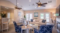 Located at 1 Hampstead Heath Way in Hampton, Virginia, this single-floor community space offers a welcoming, coastal-inspired atmosphere throughout. The open layout, visible in the dollhouse view, features a spacious L-shaped footprint that connects a comfortable lounge area with a functional kitchenette wing.

The main lounge area is styled with a relaxed coastal aesthetic, highlighted by a bold navy accent wall, a wall-mounted television, and a tiled fireplace that serves as a focal point. Plush seating, nautical-themed décor — including starfish accents and seashell artwork — and soft carpeting create an inviting gathering space. Recessed lighting and a ceiling fan keep the room bright and comfortable, while multiple sets of French doors along the perimeter flood the space with natural light and provide direct access to the outdoor surroundings.

The kitchenette is practical and well-appointed, featuring warm oak cabinetry, dark granite-style countertops, a full-size refrigerator, microwave, and a kitchen island with bar seating. Light ceramic tile flooring and a window with blinds complete the space. Coastal artwork ties the kitchenette's design back to the overall theme of the lounge, ensuring a cohesive feel throughout this well-designed community facility.