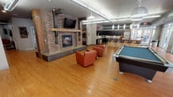 This expansive communal amenity space is designed for both relaxation and socializing, combining industrial-inspired aesthetics with warm, inviting details throughout its open single-floor layout. The centerpiece of the main lounge area is a striking full-height brick fireplace, complete with a rustic wood mantel and a wall-mounted flat-screen television above — a natural gathering point that anchors the room. Warm hardwood flooring runs throughout, complementing the earthy brick tones and contributing to the space's welcoming atmosphere. Comfortable seating, including plush leather armchairs and modular upholstered ottomans, is arranged throughout the lounge to encourage conversation and relaxation. A full-sized pool table adds a recreational dimension to the space, reinforcing its social character. A well-appointed communal kitchen sits adjacent to the main lounge, outfitted with dark espresso cabinetry, granite countertops, stainless steel appliances including a refrigerator and dishwasher, and a large central island with bar-height seating beneath sleek pendant lighting. Exposed ductwork adds an industrial touch to the kitchen area. A staircase with open wood treads and black metal railings leads to upper levels, with additional casual seating arranged at its base. The space is lit by a combination of pendant fixtures, track lighting, and recessed lighting throughout.