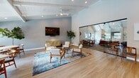 Located at 2201 Dogwood Dr in Lisle, IL, this single-floor community clubhouse combines mid-century modern aesthetics with thoughtfully designed functional spaces. The main lounge area is anchored by vaulted, white-paneled ceilings with exposed beams, recessed lighting, and a ceiling fan, creating an airy and welcoming atmosphere. A large flat-screen TV is mounted on a textured blue-gray accent wall, complemented by warm wood-framed seating arranged around a central coffee table atop a blue abstract area rug and light hardwood flooring throughout.

A glass-partition wall elegantly separates the lounge from the dining area, flooding both spaces with natural light while maintaining visual connectivity. The dining space features multiple tables with mid-century wood chairs and large black-framed windows overlooking an outdoor pool and patio area with market umbrellas.

The well-appointed kitchen includes a large white quartz island with leather bar stools, white shaker cabinetry with brass hardware, open wood floating shelves, a subway tile backsplash, and a series of gold globe pendant lights. A dedicated beverage station with a commercial coffee machine, additional counter space, and open shelving rounds out the kitchen area. Abstract wall art and abundant greenery add a refined, welcoming touch throughout the space.