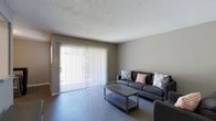 This single-floor space offers a clean and functional layout designed for comfortable everyday living. The living area serves as the centerpiece of the home, characterized by a neutral gray color palette that creates a calm and cohesive atmosphere. Large sliding glass doors stretch across one wall, allowing an abundance of natural light to fill the room and providing a seamless connection to the outdoors. Vertical blinds offer flexible light control and privacy. The living room is furnished with a contemporary dark upholstered sofa accented with patterned throw pillows, complemented by a matching low-profile coffee table that anchors the seating area. The hard surface flooring adds a sleek, low-maintenance quality to the space, while the textured ceiling adds a subtle architectural detail. The overall ambiance is understated and practical, with a modern sensibility that suits a variety of lifestyles. The open feel of the main living area, enhanced by the generous glazing and light tones, gives the space an airy and welcoming quality throughout.