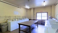 This shared laundry facility is a practical, well-organized space designed to accommodate multiple users efficiently. The room is finished with painted concrete block walls in a neutral yellow tone and features durable flooring, reflecting a straightforward, utilitarian aesthetic suited for heavy daily use.

Along one wall, a row of top-loading washing machines is neatly arranged side by side, each equipped with individual water connections and controls. Accompanying dryers are positioned adjacent, providing a complete laundry workflow within a single area. A large, centrally located folding table offers ample surface space for sorting and folding laundry, making the room highly functional. A bench near the window provides a convenient spot for waiting users.

Fluorescent light fixtures mounted on the ceiling ensure bright, even illumination throughout the space, supplemented by natural light from a large window along one wall. A posted notice board with informational signage is visible, helping occupants stay informed of rules and procedures. The overall ambiance is clean, organized, and purpose-driven, prioritizing practicality and ease of use.