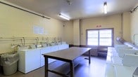 This shared laundry facility is a practical, well-organized space designed to accommodate multiple users efficiently. The room is finished with painted concrete block walls in a neutral yellow tone and features durable flooring, reflecting a straightforward, utilitarian aesthetic suited for heavy daily use.

Along one wall, a row of top-loading washing machines is neatly arranged side by side, each equipped with individual water connections and controls. Accompanying dryers are positioned adjacent, providing a complete laundry workflow within a single area. A large, centrally located folding table offers ample surface space for sorting and folding laundry, making the room highly functional. A bench near the window provides a convenient spot for waiting users.

Fluorescent light fixtures mounted on the ceiling ensure bright, even illumination throughout the space, supplemented by natural light from a large window along one wall. A posted notice board with informational signage is visible, helping occupants stay informed of rules and procedures. The overall ambiance is clean, organized, and purpose-driven, prioritizing practicality and ease of use.