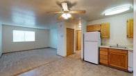 This single-floor residential space offers a straightforward and functional layout designed for comfortable everyday living. The open living area is finished with wall-to-wall carpeting and features a ceiling fan with integrated lighting, providing both comfort and illumination. A window along the exterior wall allows natural light to fill the space, contributing to a bright and airy feel.

The kitchen is positioned adjacent to the living area and is equipped with warm-toned oak cabinetry, a full-size white refrigerator, and light-colored countertops. Tile flooring in the kitchen and entryway area provides a practical, easy-to-maintain surface that transitions seamlessly from the carpeted living space.

A central hallway connects the main living and kitchen areas to the rest of the unit, with a wood-paneled interior door visible at the end, suggesting access to additional rooms. The textured ceiling and neutral wall tones throughout lend a clean, understated aesthetic. Overall, the space is designed with practicality in mind, offering a cohesive and livable environment with classic residential finishes.