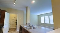 This single-floor space is designed with an open-concept layout that seamlessly connects the kitchen and living areas. The kitchen is anchored by a generous countertop with a double stainless steel sink, complemented by rich dark cabinetry beneath. A structural column adds architectural character to the space while maintaining the open flow between areas. Recessed ceiling lights and a track lighting system provide versatile illumination throughout the kitchen zone. The neutral beige and green-toned walls create a calm, welcoming ambiance across the entire floor. The adjoining living area benefits from natural light streaming through a window fitted with blinds, lending the space a bright and airy feel. Tile flooring in the kitchen transitions into the living space, offering both practicality and visual continuity. A doorway visible from the kitchen leads to a utility or laundry area, adding functional value to the layout. The overall design emphasizes clean lines, practical functionality, and a cohesive, understated aesthetic that makes the space feel organized and easy to move through.