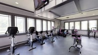 This single-floor fitness center is designed to support a comprehensive workout experience in a bright, functional environment. The main exercise area is generously proportioned, featuring elevated ceilings with a striking coffered detail that adds architectural interest and contributes to the open, airy feel of the space. A large flat-screen television is mounted overhead, keeping members entertained during their workouts.

Natural light floods the room through a series of large, multi-pane windows that line the walls, offering views of surrounding greenery and creating an inviting atmosphere. The floor is finished with durable, dark rubber gym flooring that provides both safety and practicality.

The facility is outfitted with a well-rounded selection of equipment, including recumbent and upright stationary bikes, treadmills, and a range of strength and resistance training machines. The equipment is arranged to allow comfortable circulation throughout the space. A neutral gray and white color palette, accented by touches of purple on select equipment, gives the center a clean, modern aesthetic. Overall, this fitness center offers a well-organized and comfortable environment suited for a variety of exercise routines.
