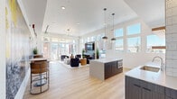 Located on Dry Creek Drive in Longmont, Colorado, this single-floor open-plan space combines a fully equipped kitchen and living area into one cohesive, light-filled environment. Light hardwood flooring runs throughout, complementing the bright white walls and elevated ceilings that lend a sense of generous openness to the interior.

The kitchen is outfitted with sleek navy cabinetry, a large white quartz island with gold-accented bar seating, illuminated open shelving against a textured white tile backsplash, a beverage refrigerator, and stainless steel appliances. Globe pendant lights hang above the island, adding a modern finishing touch. A secondary kitchen island with a built-in oven provides additional functionality.

The living area is anchored by a contemporary tile-surround fireplace topped with a wall-mounted television, framed by floor-to-ceiling windows and sliding glass doors that invite abundant natural light and outdoor views. Comfortable lounge seating, a live-edge wood coffee table, and plush area rugs create an inviting atmosphere.

A striking large-scale mural depicting Colorado outdoor adventures — skiing, rock climbing, and white-water rafting — energizes one full wall, celebrating the active lifestyle of the region. A small café-style seating nook nearby offers an intimate gathering spot within this dynamic, well-designed communal space.