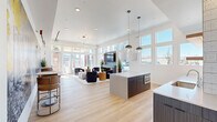 Located on Dry Creek Drive in Longmont, Colorado, this single-floor open-plan space combines a fully equipped kitchen and living area into one cohesive, light-filled environment. Light hardwood flooring runs throughout, complementing the bright white walls and elevated ceilings that lend a sense of generous openness to the interior.

The kitchen is outfitted with sleek navy cabinetry, a large white quartz island with gold-accented bar seating, illuminated open shelving against a textured white tile backsplash, a beverage refrigerator, and stainless steel appliances. Globe pendant lights hang above the island, adding a modern finishing touch. A secondary kitchen island with a built-in oven provides additional functionality.

The living area is anchored by a contemporary tile-surround fireplace topped with a wall-mounted television, framed by floor-to-ceiling windows and sliding glass doors that invite abundant natural light and outdoor views. Comfortable lounge seating, a live-edge wood coffee table, and plush area rugs create an inviting atmosphere.

A striking large-scale mural depicting Colorado outdoor adventures — skiing, rock climbing, and white-water rafting — energizes one full wall, celebrating the active lifestyle of the region. A small café-style seating nook nearby offers an intimate gathering spot within this dynamic, well-designed communal space.