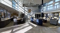This expansive single-floor space makes an immediate impression with its dramatic, soaring ceilings and an abundance of natural light streaming in through floor-to-ceiling windows that line the upper and lower walls. The open-plan layout is anchored by a bold, cylindrical pendant fireplace that serves as a striking focal point within the central lounge area. Comfortable seating is arranged thoughtfully throughout, featuring deep blue accent chairs with gold-toned legs, plush sofas, and coordinating upholstered pieces that create an inviting, upscale atmosphere. A herringbone-patterned tile floor adds texture and visual depth to the space. To one side, a dining area with a large bar-height table and upholstered chairs offers a more structured setting for gatherings. Decorative artwork and sculptural wall accents add curated character throughout. An open staircase with glass railings leads to a mezzanine-level overlook, further enhancing the sense of vertical scale. The exposed mechanical elements above blend seamlessly with the modern industrial-chic aesthetic, while lush outdoor greenery visible through the expansive glazing connects the interior to the surrounding environment.