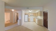 This single-floor residence offers an open and functional layout designed for comfortable everyday living. The kitchen is a central highlight, featuring light wood cabinetry that extends to the ceiling for ample storage, white appliances including a refrigerator, electric range, and dishwasher, as well as a double sink set into a continuous countertop. The kitchen flows naturally into the adjacent living area, which is finished with neutral-toned carpeting that adds warmth underfoot and contributes to the cohesive, airy feel of the space. Recessed ventilation and flush-mount ceiling lighting keep the interior bright and well-ventilated throughout. A hallway visible from the main living area leads to additional rooms, providing a clear and intuitive flow through the unit. Paneled doors and clean white walls maintain a simple, understated aesthetic throughout. The overall design emphasizes practicality and openness, making efficient use of the available floor plan while maintaining a welcoming and livable atmosphere suited to a variety of lifestyles.