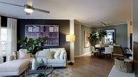 This single-floor residence showcases a thoughtfully designed open-concept layout that connects the living and dining areas in a cohesive, flowing arrangement. The living area features light hardwood flooring that extends throughout the space, complemented by a neutral-toned sofa adorned with decorative pillows and anchored by a round glass coffee table. A bold, dark accent wall serves as a striking focal point, displaying a large-scale artwork depicting rows of books, adding visual depth and a sophisticated character to the room. A ceiling fan overhead and a floor lamp provide layered lighting options. Lush indoor plants, including a large fiddle-leaf fig and tropical foliage, introduce natural elements that enhance the space's warmth and vitality. Crown molding runs along the ceiling, adding an elegant architectural detail. The dining area, visible in the background, features a round table surrounded by upholstered chairs, with track lighting above and a framed botanical artwork on the sage-toned wall. Together, these spaces create a balanced, modern aesthetic that is both functional and visually inviting.