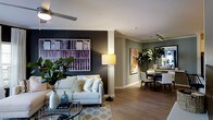 This single-floor residence showcases a thoughtfully designed open-concept layout that connects the living and dining areas in a cohesive, flowing arrangement. The living area features light hardwood flooring that extends throughout the space, complemented by a neutral-toned sofa adorned with decorative pillows and anchored by a round glass coffee table. A bold, dark accent wall serves as a striking focal point, displaying a large-scale artwork depicting rows of books, adding visual depth and a sophisticated character to the room. A ceiling fan overhead and a floor lamp provide layered lighting options. Lush indoor plants, including a large fiddle-leaf fig and tropical foliage, introduce natural elements that enhance the space's warmth and vitality. Crown molding runs along the ceiling, adding an elegant architectural detail. The dining area, visible in the background, features a round table surrounded by upholstered chairs, with track lighting above and a framed botanical artwork on the sage-toned wall. Together, these spaces create a balanced, modern aesthetic that is both functional and visually inviting.