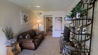 This single-floor space features a living room with a warm, inviting ambiance defined by neutral beige walls and wall-to-wall carpeting that adds a sense of comfort underfoot. The textured ceiling with crown molding detailing lends a classic, finished look to the room. A plush brown leather sofa serves as the centerpiece of the living area, complemented by decorative accent pillows, while a floor lamp provides warm ambient lighting. An ornate wrought-iron shelving unit on one side of the room offers display space for plants and decorative items, adding visual interest and a touch of greenery to the interior. Wall art, including a large leaf-motif canvas, contributes to the room's tasteful, casual aesthetic. A flat-screen television is positioned for comfortable viewing. An open doorway leads to an adjacent room, suggesting a connected and functional layout throughout the space. The overall atmosphere is cozy and well-appointed, with thoughtful décor choices that create a lived-in, homely feel suited for everyday relaxation.