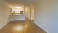 This single-floor residence offers a clean, open-concept layout that seamlessly connects the living and kitchen areas. Warm hardwood flooring extends throughout the main living space, adding a sense of continuity and comfort underfoot. Classic crown molding lines the ceilings, lending an elegant, traditional touch to the interior.

The kitchen is defined by white cabinetry and stainless steel appliances, including a refrigerator and range, with a raised breakfast bar providing both a functional workspace and a casual dining option. Pendant lighting above the bar adds a stylish focal point to the kitchen zone.

Decorative columns with white trim frame the transition between the kitchen and the open living area, reinforcing the architectural character of the space. The neutral, warm-toned walls create a bright and inviting atmosphere throughout.

A hallway leading from the main living area provides access to additional rooms, including what appears to be a closet or utility space accessible through a paneled door. The layout is efficient and practical, making good use of the available floor plan. Overall, the space presents a well-maintained, move-in ready interior with timeless design elements suitable for everyday living.