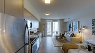 This single-floor space showcases a thoughtfully designed open-concept layout that seamlessly connects the kitchen and living areas. The kitchen is positioned along one wall and features stainless steel appliances, including a refrigerator and range, complemented by sleek cabinetry with a warm wood-tone finish and a white tile backsplash. Ample upper and lower cabinet storage provides generous functionality.

The living area extends naturally from the kitchen, finished with light hardwood-style flooring throughout that contributes to a cohesive, contemporary aesthetic. Flush-mount ceiling lighting illuminates both zones evenly. Neutral-toned walls and sheer floor-to-ceiling curtains enhance the sense of openness and allow natural light to filter through generously.

A standout feature is the glass-panel door at the far end of the living space, which opens directly onto a private balcony with views of the surrounding urban skyline. The overall ambiance is modern and refined, with a neutral palette accented by gold and warm tones in the furnishings, creating a welcoming and stylish environment suited to both relaxing and entertaining.