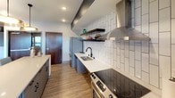 This contemporary single-floor space is designed with an open-concept layout that seamlessly connects the kitchen, dining, and lounge areas. The kitchen is outfitted with stainless steel appliances — including a French door refrigerator, electric range, and dishwasher — complemented by sleek quartz countertops, a matte black faucet, and a striking vertical white tile backsplash. Open floating wood shelves add a functional yet stylish touch, while a stainless steel wall-mounted range hood and pendant lighting enhance the modern aesthetic. Wood-look flooring runs throughout, providing warmth and continuity across the space.

The dining area features a solid wood table surrounded by upholstered seating, anchored by a bold circular halo pendant light. A patterned accent wall and a window offering outside views bring personality and natural light to the space.

The lounge area is a standout, centered around a floor-to-ceiling dark tile fireplace wall with a mounted flat-screen television. A coffered ceiling with globe pendant lights defines the zone, while built-in banquette seating with a geometric accent wall creates an intimate nook. A comfortable sectional sofa and layered area rugs complete the inviting atmosphere. Together, these elements create a cohesive, well-appointed space ideal for both casual gatherings and relaxed everyday living.