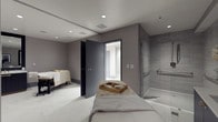 This single-level spa suite offers a serene and professionally appointed environment designed for therapeutic treatments and relaxation. The space features two massage tables dressed in clean, neutral linens, each accompanied by amenity trays stocked with treatment essentials. The interior is finished in a sophisticated palette of soft grays and warm whites, creating a calm and clinical ambiance well-suited for wellness services.

Recessed ceiling lighting provides even, unobtrusive illumination throughout the suite, complementing the smooth light-toned flooring that runs seamlessly across the space. A glass-enclosed walk-in shower with gray tile surround and integrated grab bars occupies one corner of the room, offering practical post-treatment amenities. An attached bathroom, visible through a dark-framed door, adds further convenience for guests.

The layout is efficiently organized, allowing clear separation between treatment and wet areas while maintaining an open, uncluttered feel. The neutral wall tones and clean architectural lines contribute to the overall sense of quiet professionalism, making this suite well-suited for a range of body and massage treatments in a contemporary wellness setting.