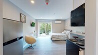 This single-floor studio unit offers a well-designed open-concept layout that efficiently combines living, sleeping, and kitchen functions within one bright, cohesive space. Light grey laminate flooring runs throughout, complementing the soft neutral wall tones and white trim to create a clean, modern aesthetic.

The main living and sleeping area is anchored by a full-sized bed and benefits from generous natural light through a large sliding glass door that opens to an outdoor view of mature trees, lending the space a calm, nature-connected ambiance. A wall-mounted television, a compact console table, and a modern egg-style swivel chair complete the living arrangement. A wall-mounted mini-split system provides efficient climate control.

The kitchen is neatly tucked along one wall and features granite countertops, white shaker cabinetry, a stainless steel electric range with an overhead microwave, a refrigerator, and a corner sink — all the essentials for full meal preparation. Built-in open shelving along the entry corridor provides additional storage.

The private bathroom is finished with matching granite countertops on a white vanity, a glass-enclosed shower, and wood-toned flooring, maintaining the unit's consistent, polished design language throughout.