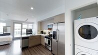 This single-floor space features a thoughtfully designed kitchen that serves as a central highlight. The kitchen showcases stainless steel appliances, including a French door refrigerator with a water and ice dispenser, a gas range, a built-in microwave, and a dishwasher. Light-toned, flat-front cabinetry extends to the ceiling, maximizing storage while contributing to a clean, contemporary aesthetic. A neutral tile backsplash adds subtle texture between the upper and lower cabinets. The countertops are finished in a light, solid surface material that complements the overall color palette. A stainless steel sink is positioned beneath a window that allows natural light to fill the space. Dark hardwood-style flooring grounds the room and provides a warm contrast to the lighter cabinetry and walls. Recessed and track lighting ensure the kitchen is well-lit. A partially visible laundry area is accessible just off the kitchen, featuring stacked washer and dryer units. The open sightline from the kitchen toward what appears to be a living area enhances the sense of flow and connectivity throughout the single-floor layout.