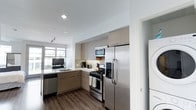 This single-floor space features a thoughtfully designed kitchen that serves as a central highlight. The kitchen showcases stainless steel appliances, including a French door refrigerator with a water and ice dispenser, a gas range, a built-in microwave, and a dishwasher. Light-toned, flat-front cabinetry extends to the ceiling, maximizing storage while contributing to a clean, contemporary aesthetic. A neutral tile backsplash adds subtle texture between the upper and lower cabinets. The countertops are finished in a light, solid surface material that complements the overall color palette. A stainless steel sink is positioned beneath a window that allows natural light to fill the space. Dark hardwood-style flooring grounds the room and provides a warm contrast to the lighter cabinetry and walls. Recessed and track lighting ensure the kitchen is well-lit. A partially visible laundry area is accessible just off the kitchen, featuring stacked washer and dryer units. The open sightline from the kitchen toward what appears to be a living area enhances the sense of flow and connectivity throughout the single-floor layout.