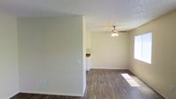 This single-story space offers a straightforward, functional layout with a clean and neutral aesthetic throughout. The main living area features wood-look plank flooring in a warm medium-brown tone, providing a durable and easy-to-maintain surface underfoot. Walls are finished in a soft, light neutral tone that keeps the interior feeling bright and open, complemented by white baseboards that add a crisp contrast.

The textured popcorn ceiling adds a classic character to the space, while a ceiling fan with a light fixture is centrally positioned in the main room, offering both air circulation and ambient lighting. A window along one wall allows natural light to filter into the room, casting soft light across the flooring.

Partially visible from the main living area is what appears to be a kitchen space, distinguished by white cabinetry and a countertop with a darker finish. A partial wall separates the two areas, providing a degree of definition between the living and kitchen zones while maintaining an open connection between them. Overall, the space presents a simple, move-in-ready environment with practical finishes suited for everyday living.