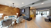 This expansive single-floor amenity space is designed with a modern, upscale aesthetic that seamlessly blends entertainment and relaxation. The open-concept layout flows between distinct zones, each thoughtfully appointed with contemporary finishes and furnishings throughout.

The game room area serves as a centerpiece, featuring a sleek ping-pong table and a full-size billiards table positioned beneath stylish pendant lighting. A bold geometric gold-toned wall sculpture adds a striking decorative focal point nearby. Adjacent to the game area, a lounge zone is anchored by a linear gas fireplace set within a textured dark stone surround, complemented by a warm wood accent wall and plush seating with eclectic accent chairs.

A well-equipped wet bar runs along one wall, offering marble-topped dark cabinetry, built-in beverage coolers, a microwave, and a wall-mounted television above a glossy white subway tile backsplash. A raised bar counter with seating provides an additional gathering spot nearby.

Wood-look tile flooring unifies the space throughout, while recessed lighting keeps the interior bright. Large glass doors along one wall flood the space with natural light and open directly to an outdoor pool courtyard, creating a seamless indoor-outdoor connection.