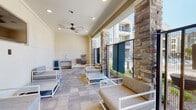This covered outdoor lounge is a well-appointed communal amenity space designed for both relaxation and convenience. The area is defined by striking natural stone columns that frame open sightlines to the surrounding courtyard and multi-story residential buildings beyond. Underfoot, large-format tile pavers in a neutral tone extend throughout the space, lending a clean and cohesive aesthetic.

Multiple seating arrangements composed of white-framed chairs and loveseats with gray upholstered cushions are thoughtfully positioned throughout, providing ample room for gathering. Overhead, ceiling fans are mounted to the covered ceiling, keeping the space comfortable year-round, while recessed lighting ensures usability into the evening hours. A wall-mounted flat-screen television is installed on one end, adding an entertainment element to the lounge.

A dedicated utility station along one wall features a stainless steel utility sink alongside a built-in water fountain, offering practical conveniences for residents. At the far end of the lounge, large glass doors and windows open to a fitness center, which is visible and directly accessible from the lounge, housing a variety of cardio and exercise equipment. The overall design blends indoor comfort with outdoor openness, creating a versatile, community-focused environment.