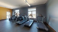 This single-level fitness center is thoughtfully designed to support a full range of workout routines in a clean, professional environment. The space is divided into distinct functional zones, each tailored to a specific type of exercise.

The cardio area features multiple treadmills and elliptical machines arranged along a wall of windows, offering views of the outdoors and filling the space with natural light. The cool blue-gray walls and durable rubber flooring give this zone a focused, energetic atmosphere.

Adjacent to the cardio section is a strength training area with a full rack of dumbbells in graduated sizes, a cable machine, a weight bench, and a wall mirror — providing everything needed for resistance training. A wooden bench adds a practical touch for rest or gear storage.

A dedicated cycling studio, set apart with warm golden-yellow walls, houses multiple stationary spin bikes, medicine balls, resistance bands, and a wall-mounted flat-screen television, creating an immersive group or solo cycling experience. Stylish pendant lighting enhances the ambiance of this room.

The space also includes a functional training zone with a rowing machine, a captain's chair, and exercise mats, bathed in natural light from large windows. Throughout, the rubber-speckled flooring provides durability and safety, while the combination of blue and gold tones creates a cohesive and motivating environment.