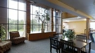 This single-floor space makes a striking architectural statement through its expansive floor-to-ceiling window wall, which floods the interior with abundant natural light and frames lush outdoor greenery. The façade combines warm brick columns with large metal-framed glass panels, creating a seamless visual connection between the indoors and the surrounding landscape. Interior potted trees placed along the window ledge reinforce this organic, nature-forward aesthetic. The dining area is thoughtfully appointed with dark wood tables and matching slatted chairs, offering ample seating for communal gatherings. Decorative greenery on the tables adds a fresh, inviting touch. The neutral carpet flooring and soft, ambient lighting from ceiling-mounted fixtures contribute to a calm and comfortable atmosphere throughout. An open view into an adjacent kitchen area — featuring white cabinetry and a functional layout — suggests that this space is well-suited for organized meal service or community dining. The overall design blends functional architecture with warm, natural materials, resulting in a space that is both practical and aesthetically welcoming for its occupants and visitors alike.